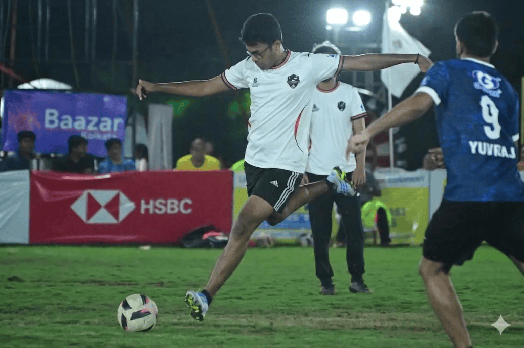 A wide-angle view of a grassroots football match on a Kolkata neighbourhood ground, players in motion under bright floodlights with the city's rooftops visible in the background.